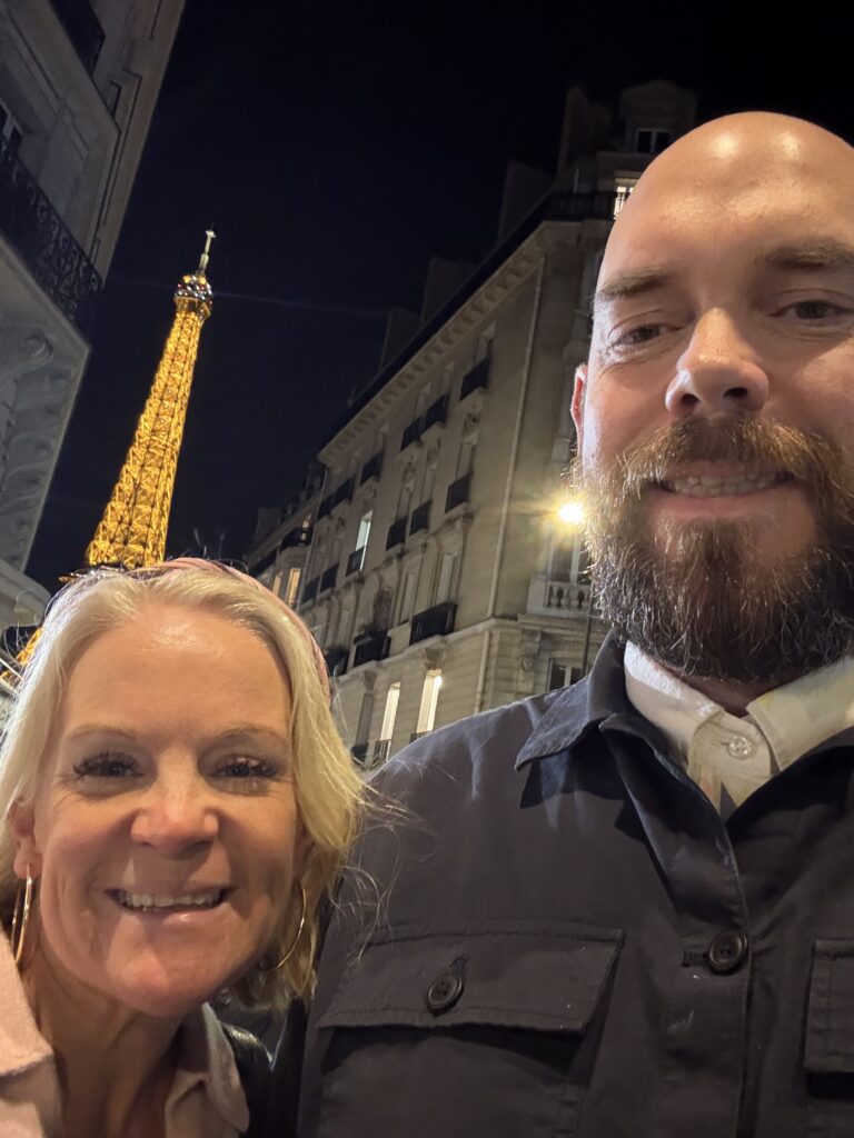 Issy and her husband smiling in a Paris street selfie with the illuminated Eiffel Tower behind them