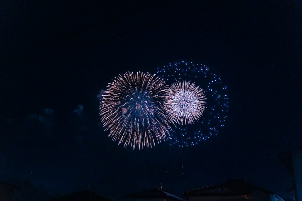 Two fireworks bursting in a dark night sky above rooftops